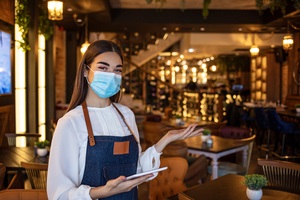 happy waitress working at a restaurant wearing a facemask and using a digital tablet and looking at the camera smiling