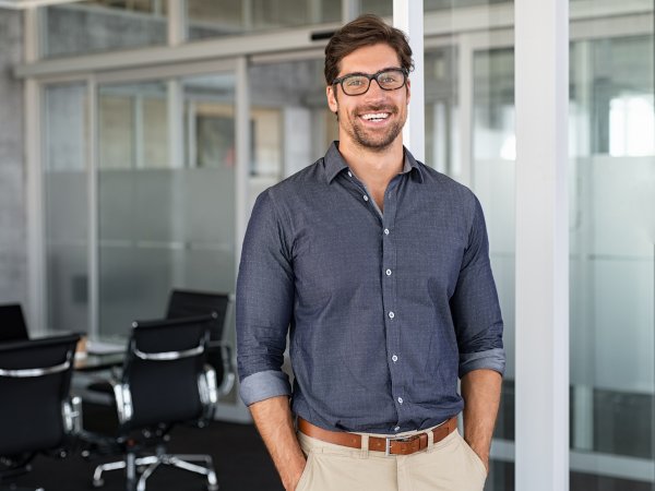 Portrait of young businessman wearing eyeglasses and standing outside conference room. Portrait of happy business man wearing spectacles and looking at camera with copy space. Satisfied proud man feeling confident in a modern office.