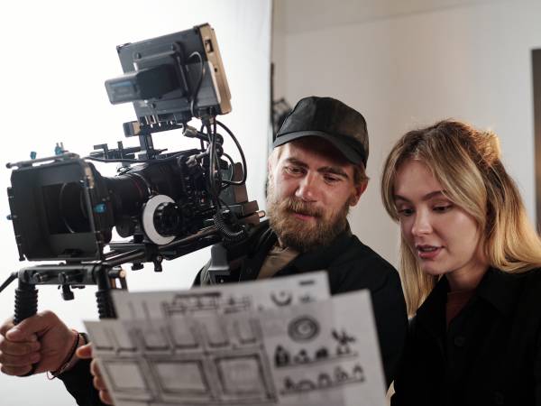 Bearded videographer with videocamers and his blond female assistant discussing working documents during studio shooting