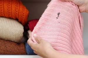 woman hands holding woolen knitted cloth with hole made by moth over wardrobe with stacks cloth on shelf